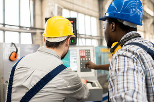 Rear view of serious busy young multiethnic factory employees in hardhats discussing program while working with control panel of powerful automated machine